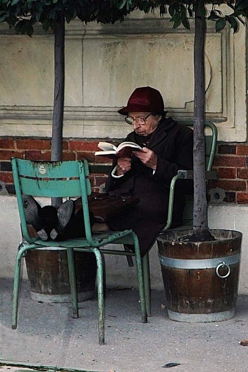 an old woman sitting on a chair reading a book next to a tree and a barrel