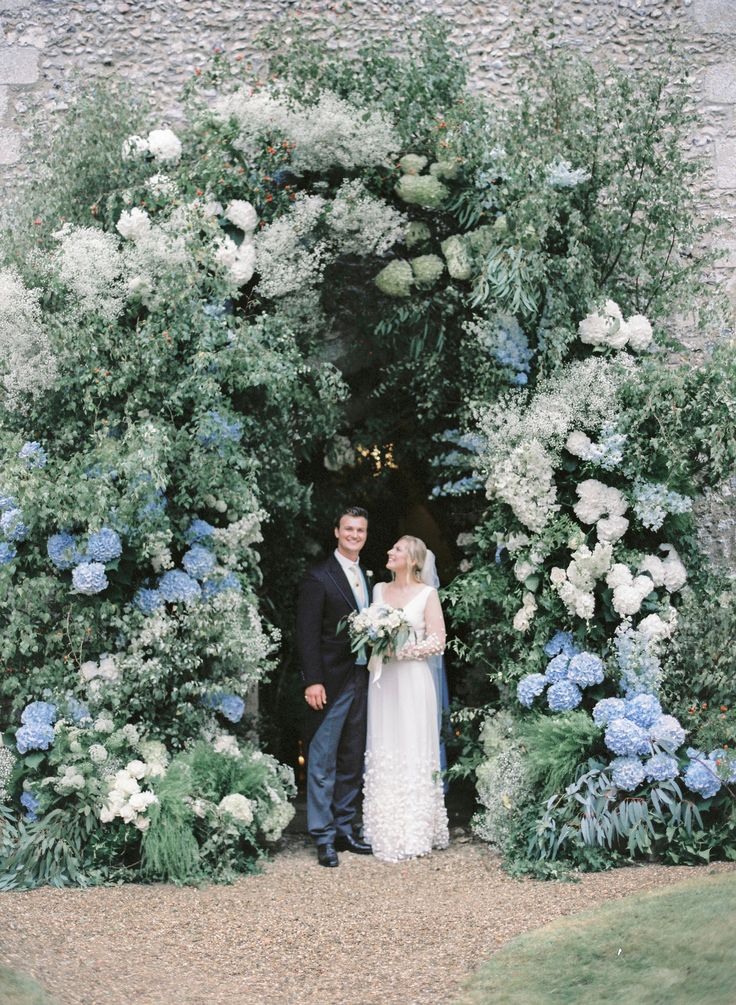 a bride and groom standing in front of a floral arch at their wedding ceremony, surrounded by blue and white flowers