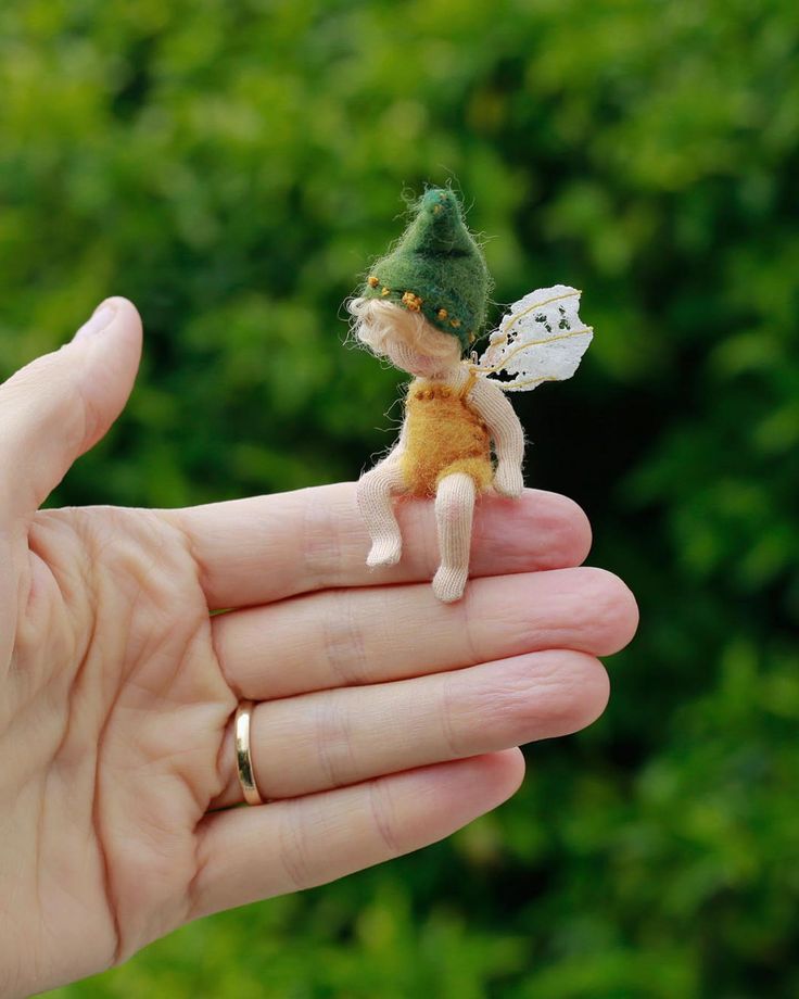 a tiny toy fairy sitting on top of someone's hand with trees in the background