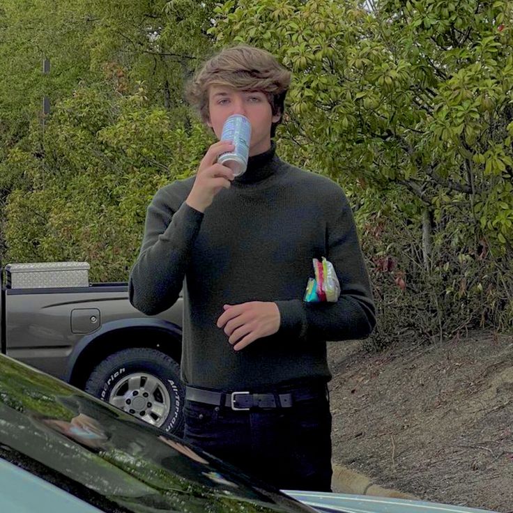 a young man drinking from a can while standing next to a car