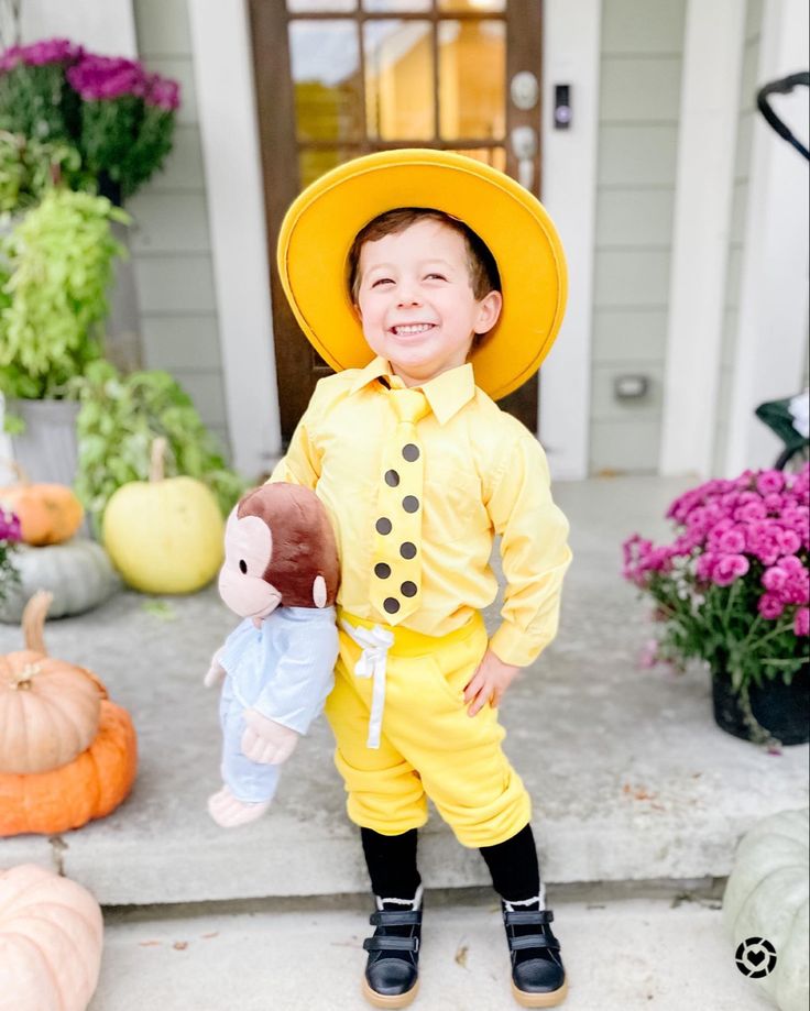 a little boy in a yellow outfit and hat holding a teddy bear on the front porch