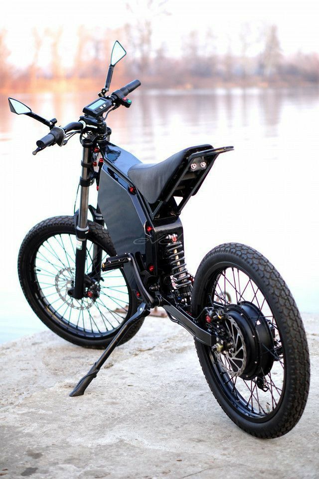 a black motorcycle parked on top of a sandy beach next to water and trees in the background