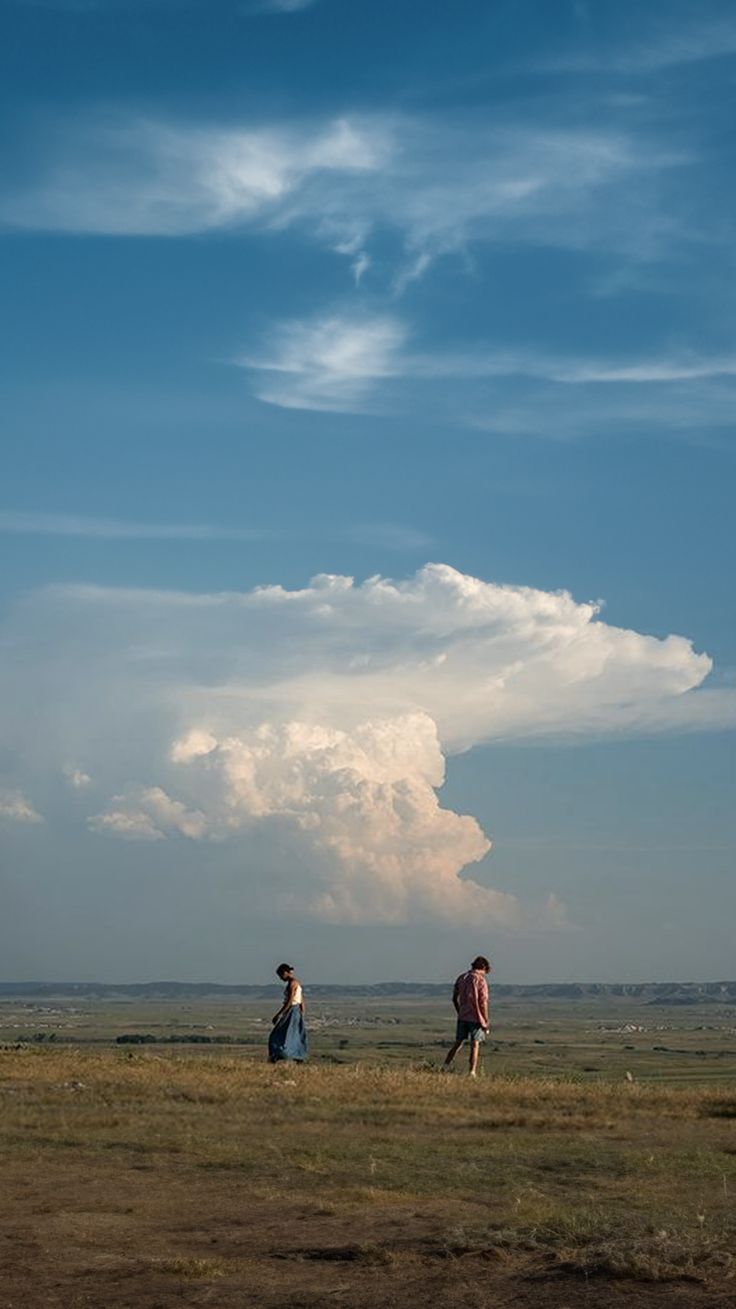 two people walking in an open field under a blue sky with white clouds above them