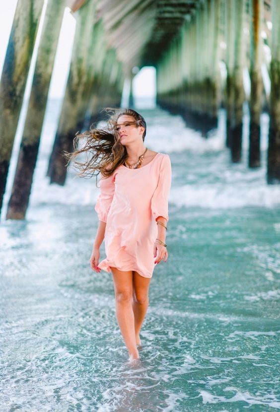 a woman is walking in the water under a pier with her hair blowing in the wind