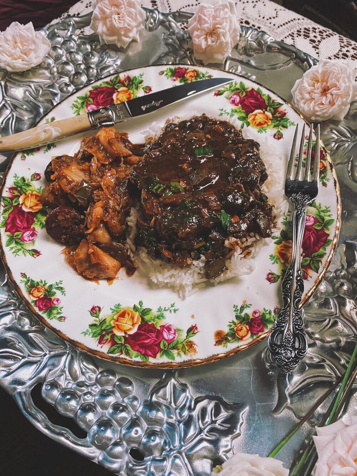 a plate with meat and rice on it next to silver utensils, flowers