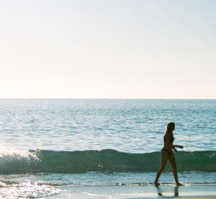 a woman is walking along the beach with her surfboard in hand and water behind her