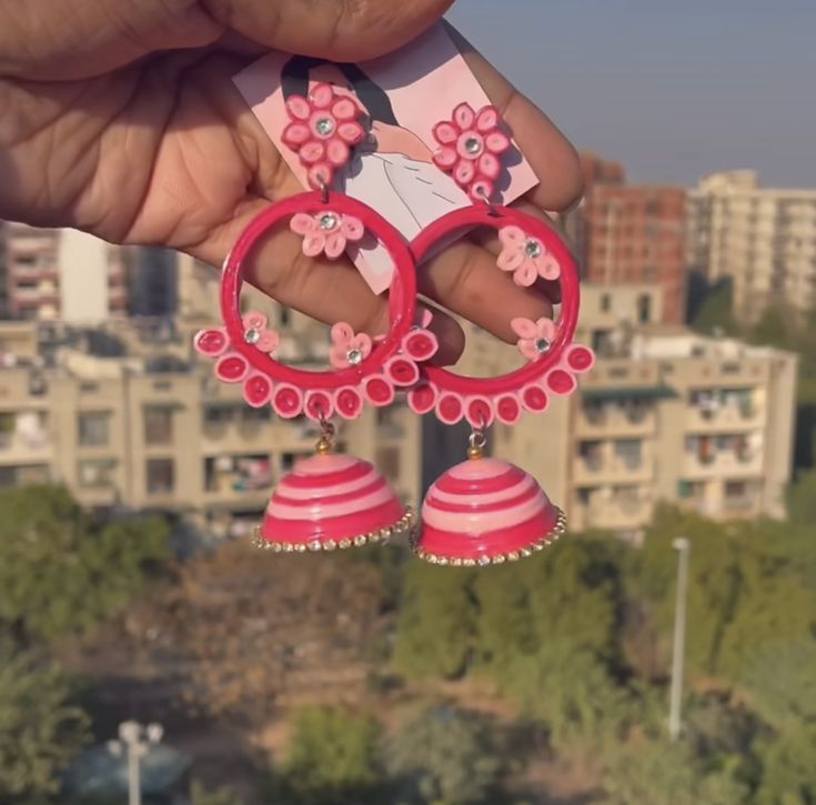 a person holding up some pink earrings in front of a cityscape with buildings