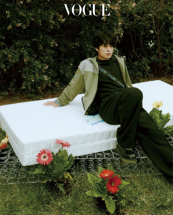 a young man sitting on top of a mattress in the middle of some grass and flowers