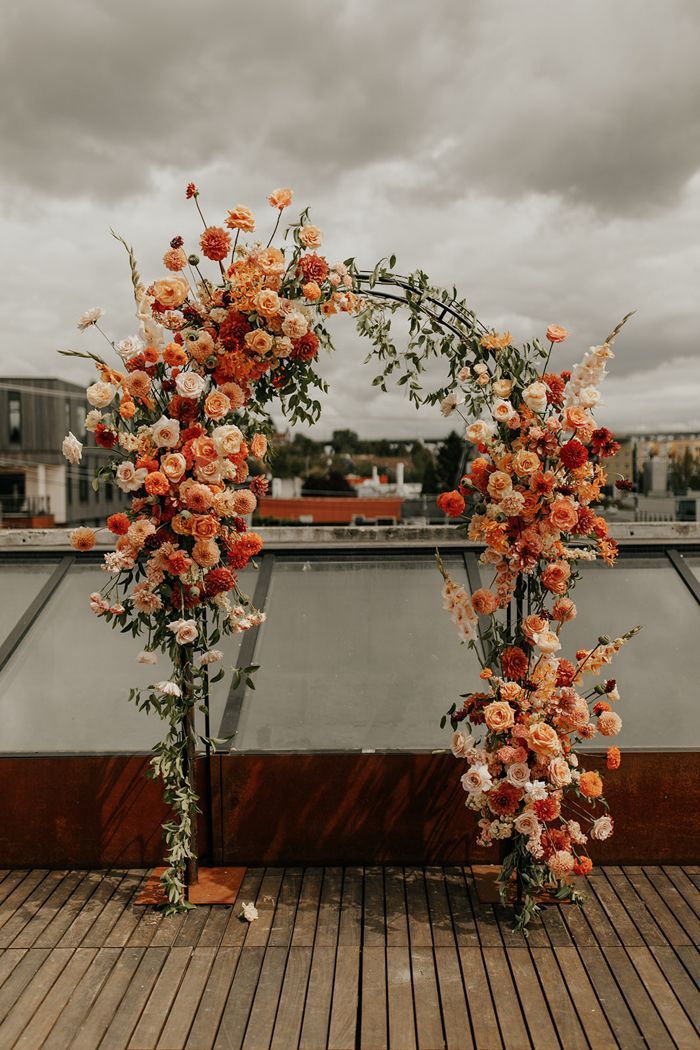 an arch decorated with orange and white flowers on top of a wooden floor next to a body of water