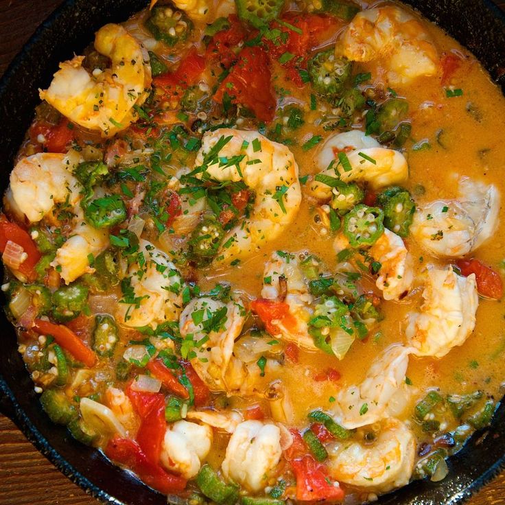 a skillet filled with shrimp and vegetables on top of a wooden table next to a spoon