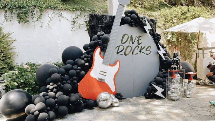 a rock and roll sign is surrounded by balloons in the shape of guitars, with people sitting around it