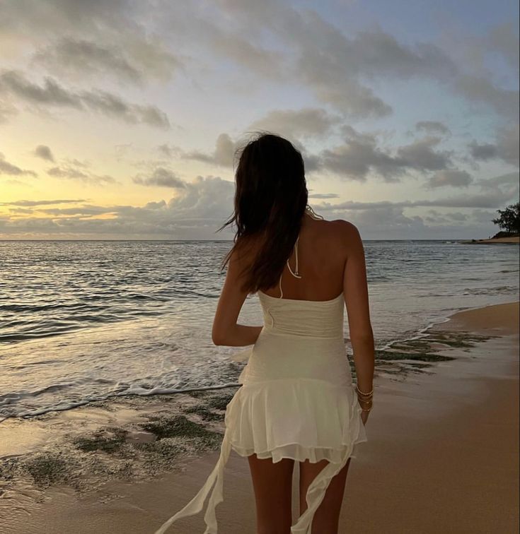 a woman in a white dress walking on the beach at sunset with her back to the camera