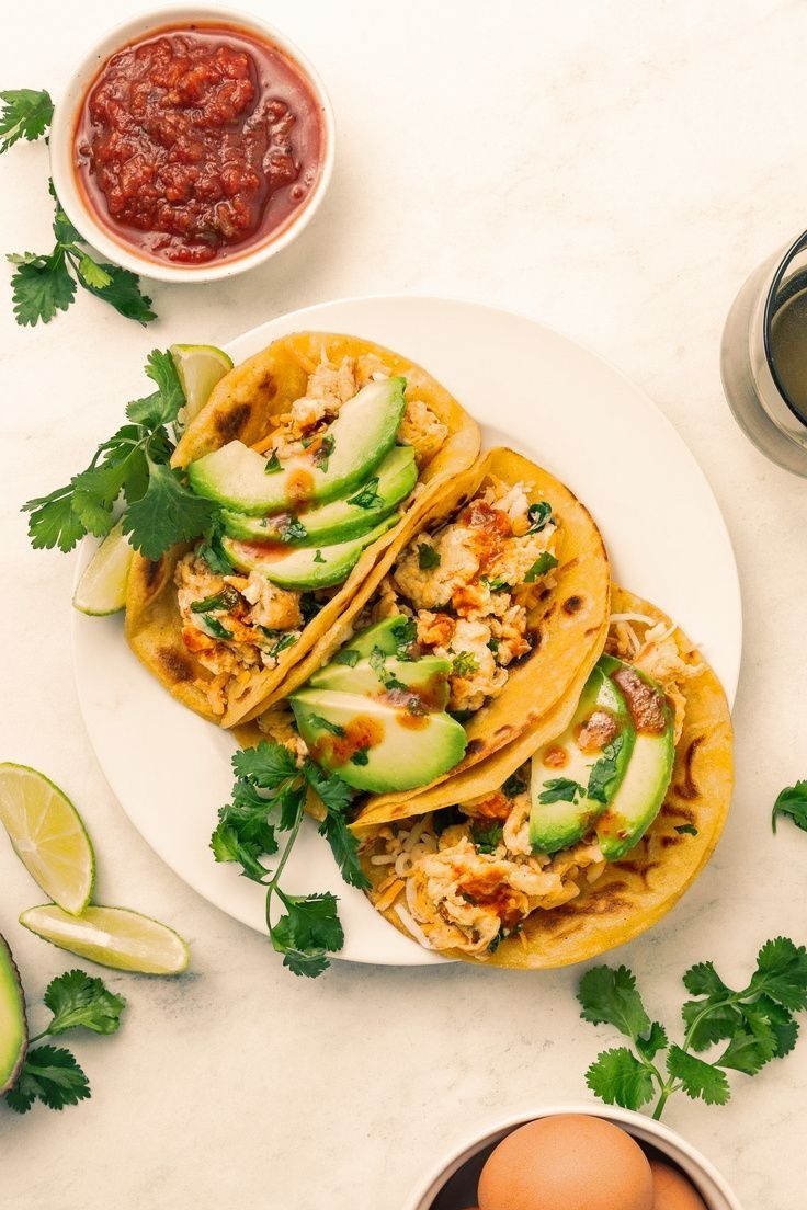 three tacos on a white plate with salsa and avocado next to it