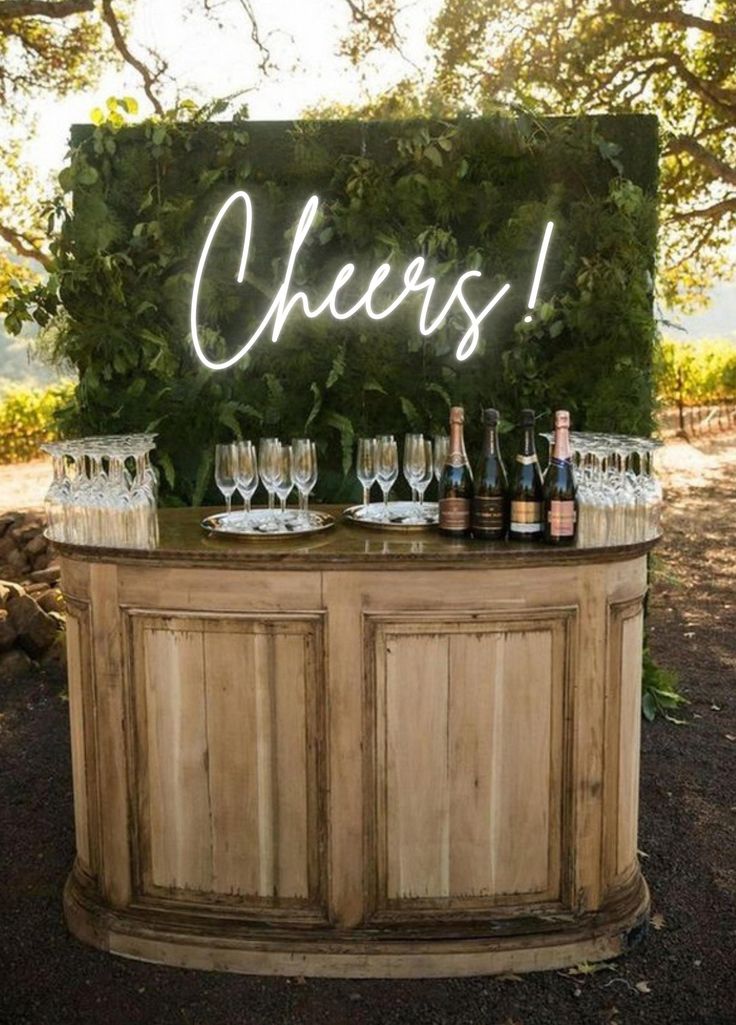 an outdoor bar with wine glasses and bottles on the top, under a neon sign that says cheers