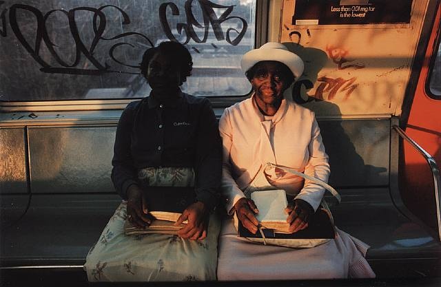 two women sitting on a bench in front of a window with graffiti written on it