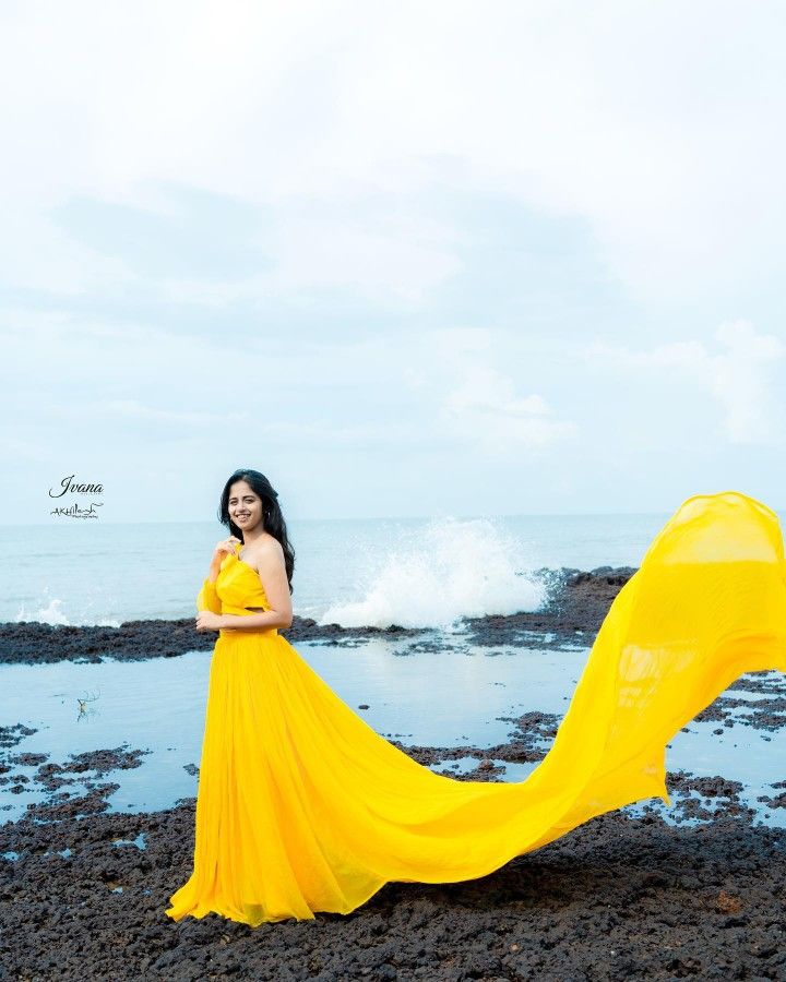 a woman in a yellow dress is standing on the rocks by the ocean with her long flowing fabric