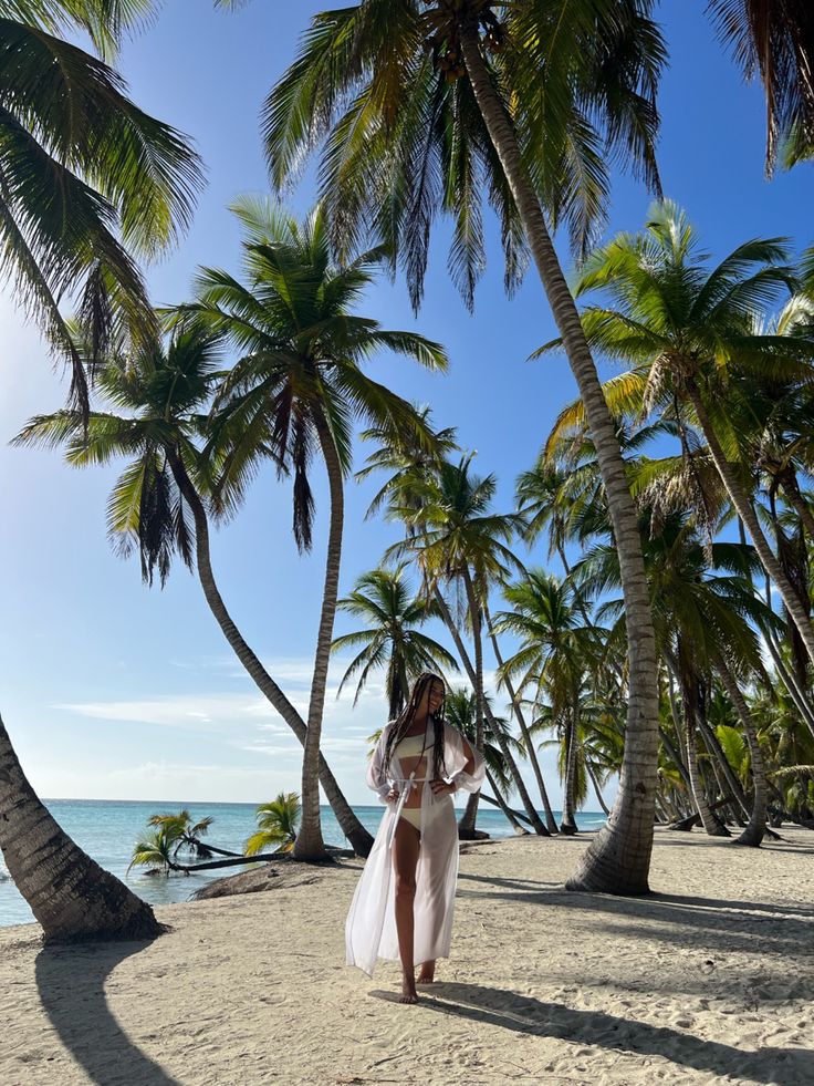 a woman in a white dress walking on the beach with palm trees and blue water