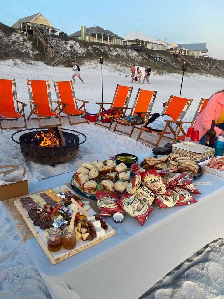 a table full of food on the beach next to chairs and fire pit with people in the background