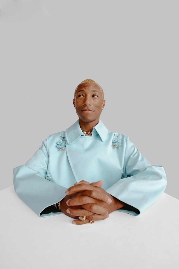 a man sitting at a table with his hands folded in front of him, wearing a blue shirt and gold bracelet