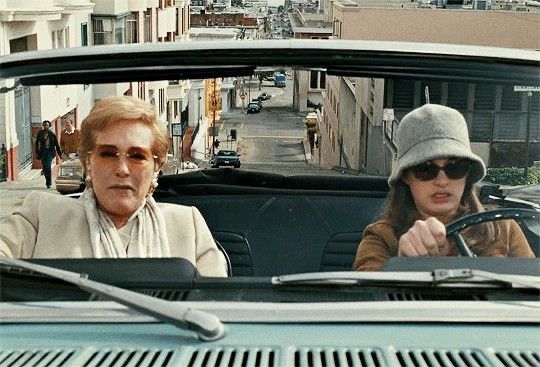 two women sitting in the driver's seat of an old car on a city street