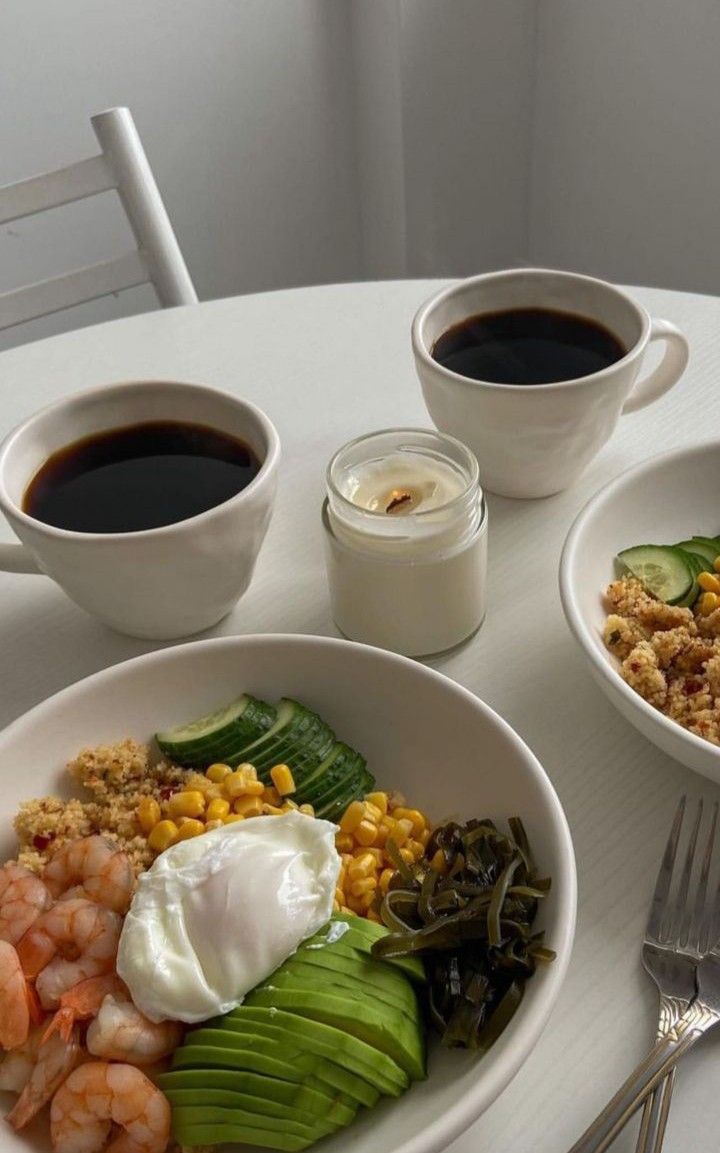 two bowls of food are sitting on a table next to cups of coffee and spoons