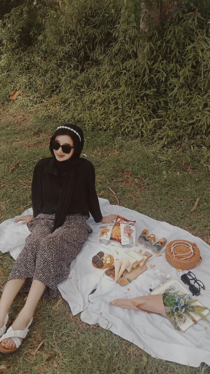 a woman sitting on top of a blanket in the grass next to some food and drinks