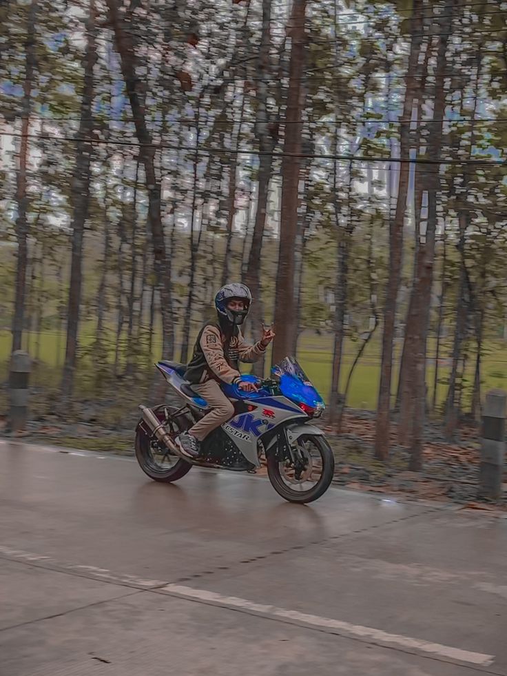 a man riding a motorcycle down a street next to tall trees and grass on a rainy day