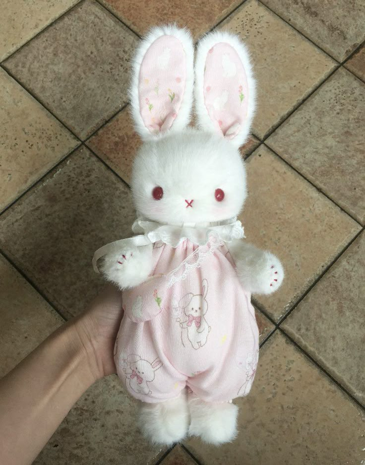 a hand holding a stuffed animal bunny in pink clothing on the ground with tile flooring