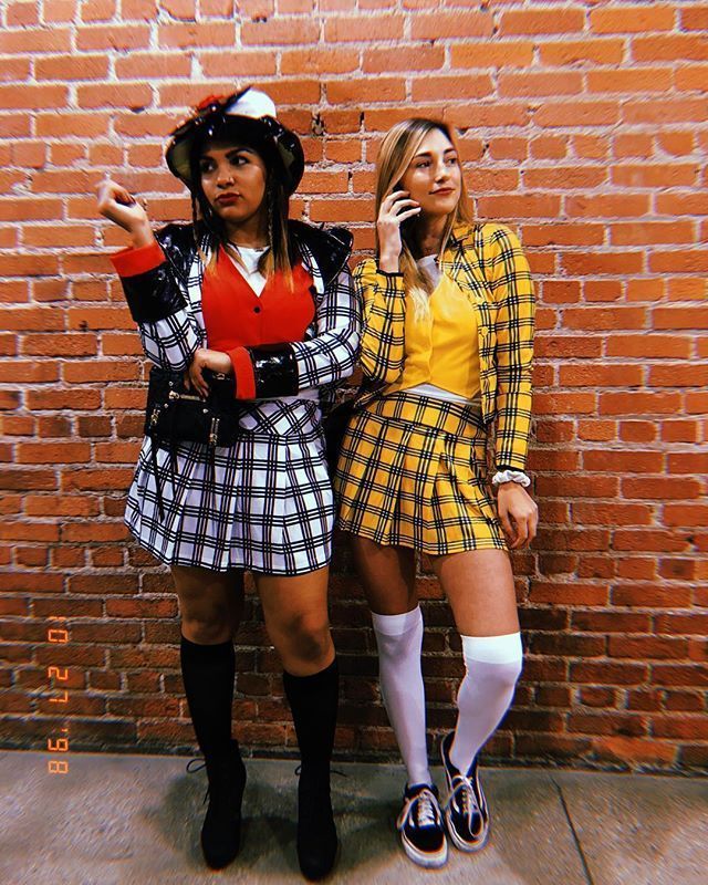 two women dressed in costumes standing next to a brick wall talking on their cell phones