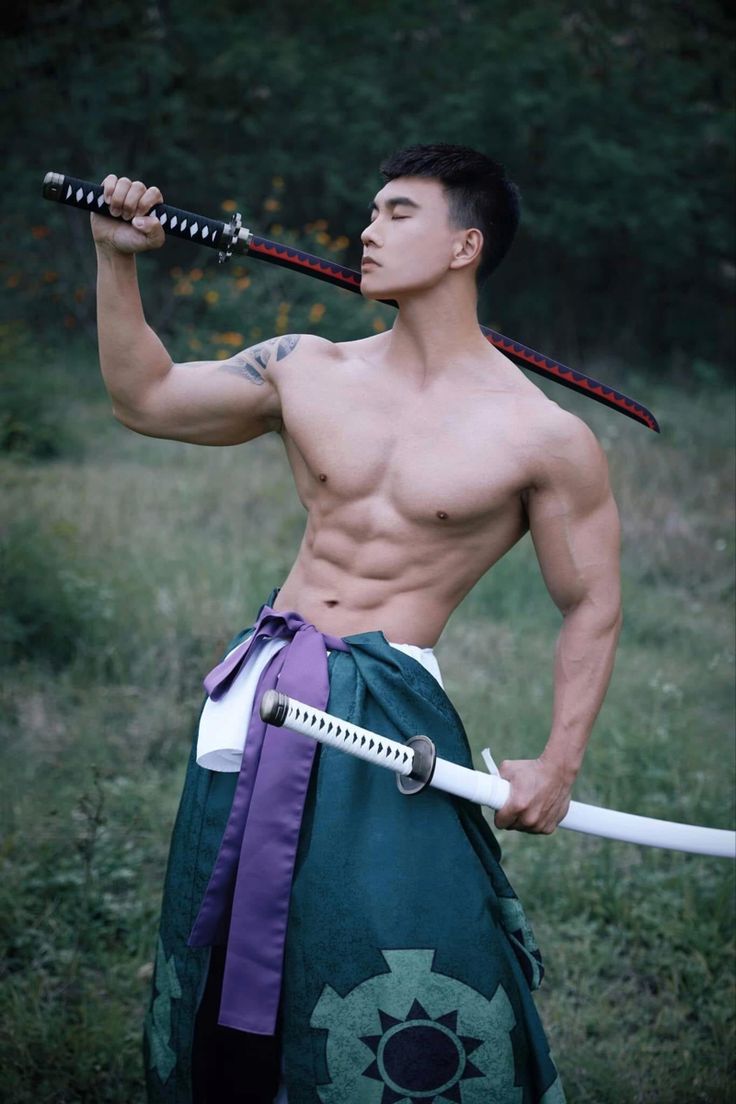 a shirtless man holding two swords while standing in the grass with trees behind him