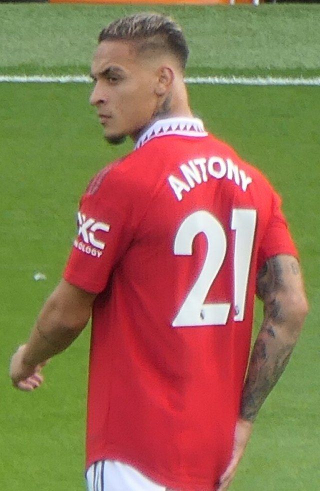 a man with tattoos on his arm standing in front of a soccer field wearing a red jersey