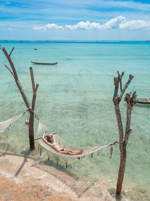 a man laying in a hammock on the beach next to some trees and boats