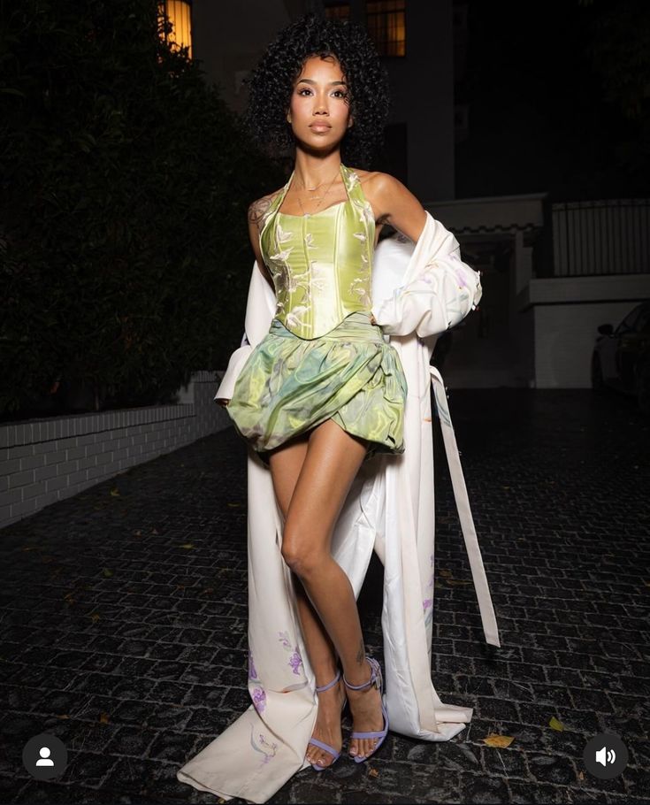 a woman in a green dress and white coat posing for the camera with her hands on her hips