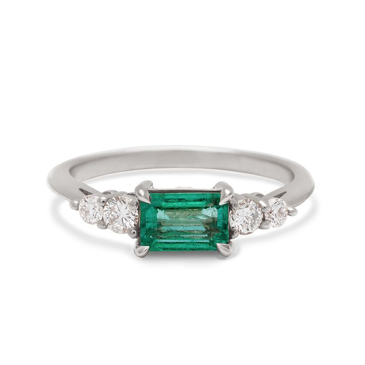 A ring with an emerald cut and shaped green emerald center stone with two round white diamonds on either side set in white gold. 14k White Gold Emerald Ring With Brilliant Cut, Gia Certified Emerald Cut Diamond Ring, Emerald Ring With Center Stone And Emerald Cut, Timeless Emerald Cut May Birthstone Rings, Classic 14k White Gold Emerald Ring For May Birthstone, Gia Certified Emerald Cut Emerald Ring For May Birthstone, White Gold Emerald-cut Emerald Ring, White Gold Emerald Cut Emerald Ring, Emerald Cut Emerald Ring With Center Stone