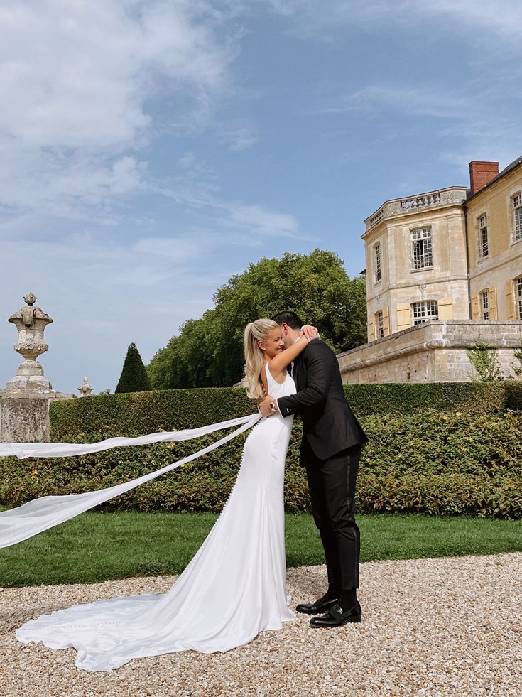 a bride and groom kissing in front of a large building with hedges on the side