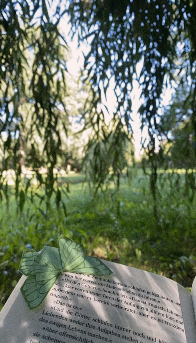 an open book sitting on top of a lush green field next to a leaf covered tree