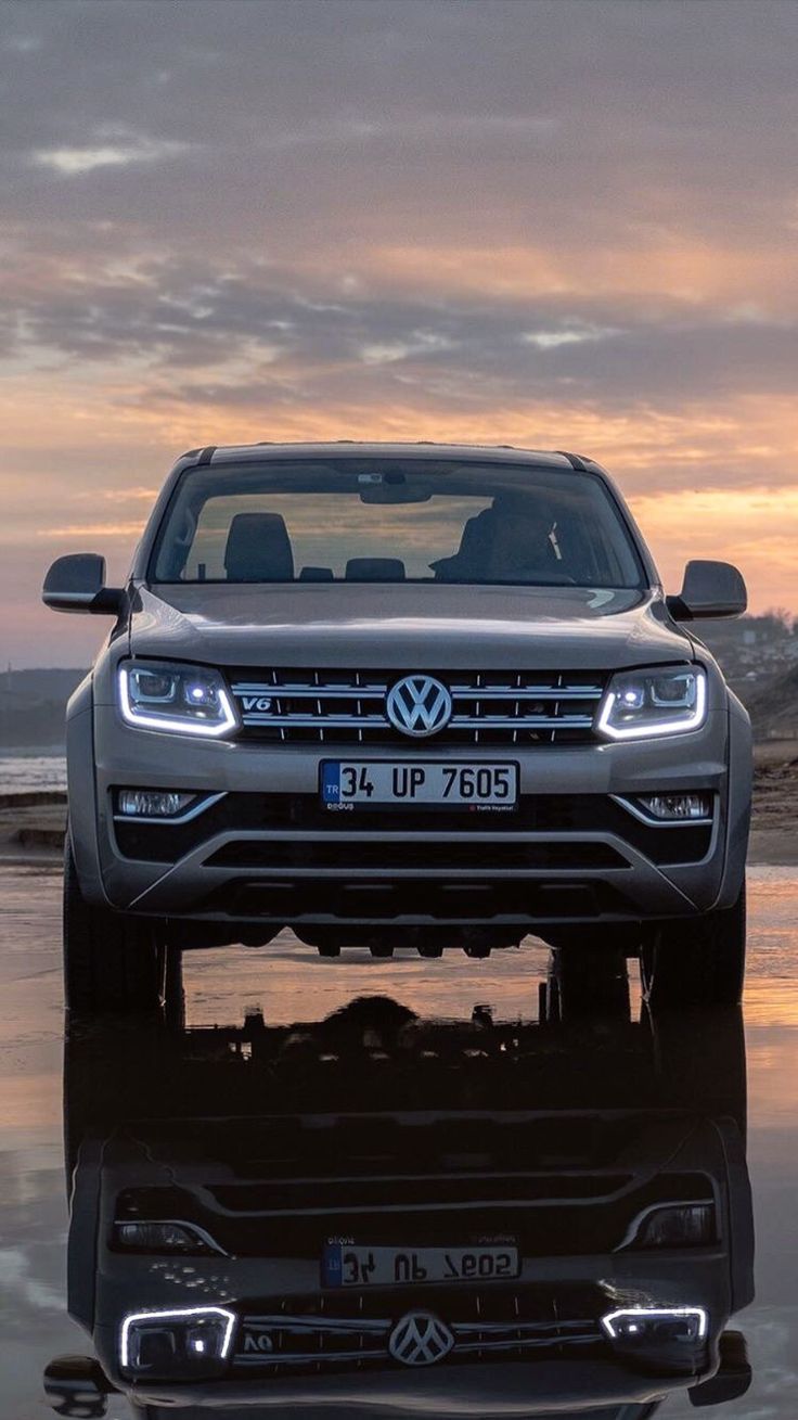 the front end of a silver volkswagen suv parked on top of a wet beach at sunset