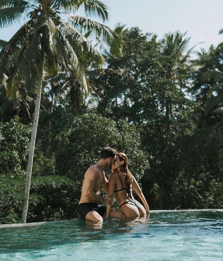 a man and woman sitting on the edge of a swimming pool next to palm trees