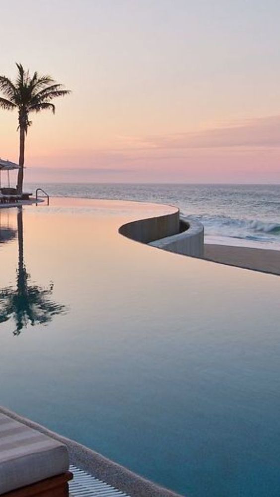 an empty swimming pool next to the ocean at sunset with palm trees in the foreground