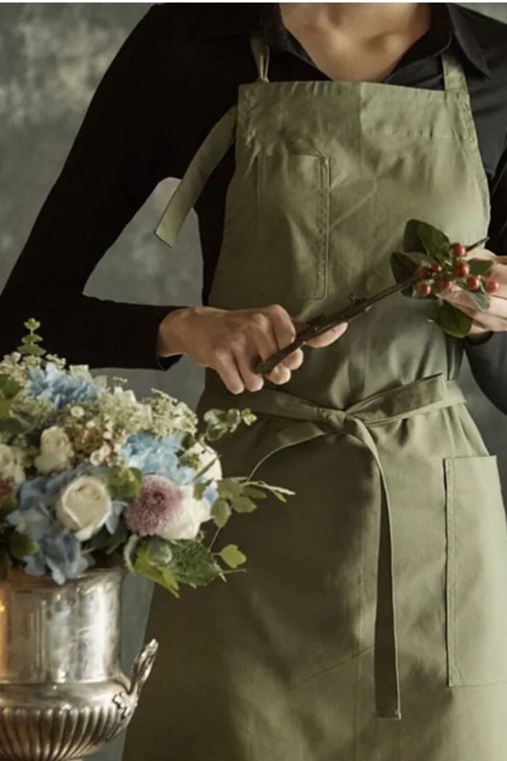 a woman in an apron is holding a knife and flower arrangement while standing next to a vase with flowers