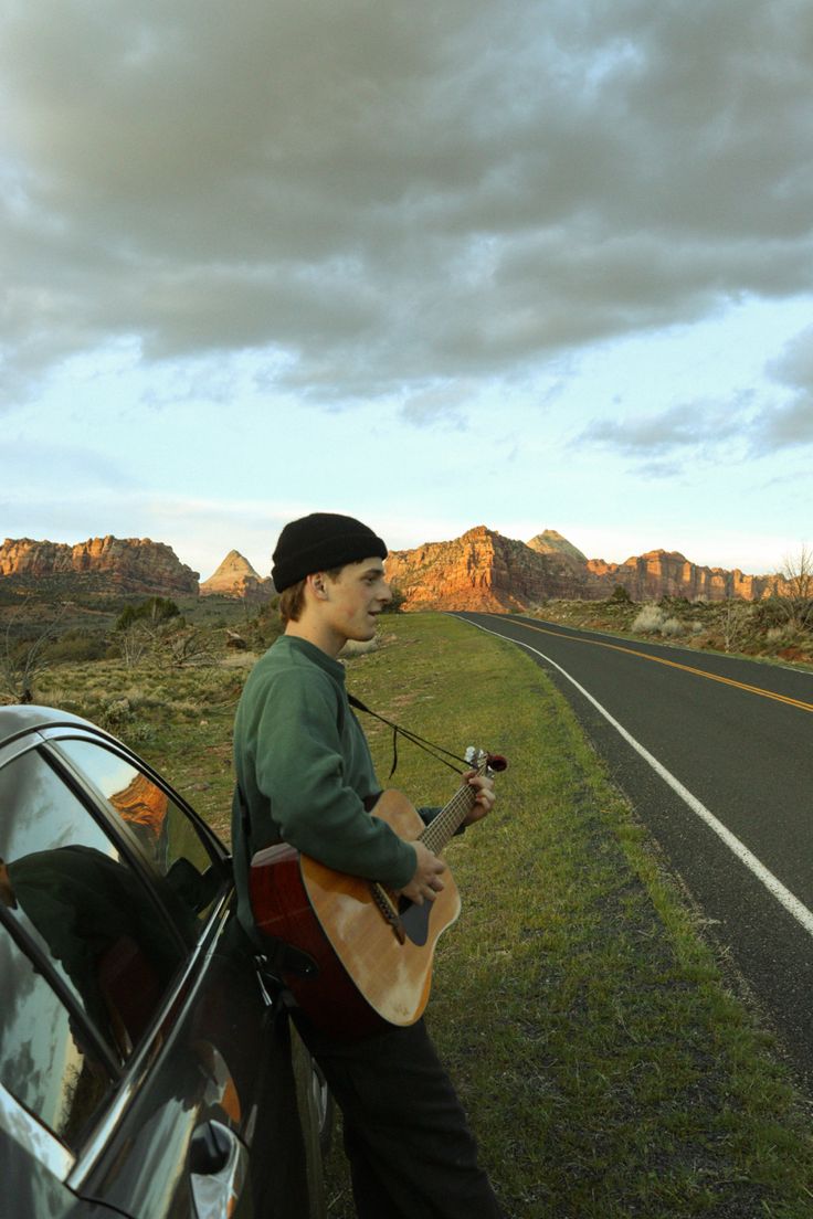 a man with a guitar standing next to a car on the side of the road