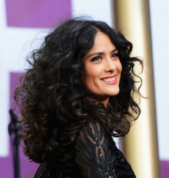 a woman with curly hair smiling and wearing a black lace dress, standing in front of a purple background