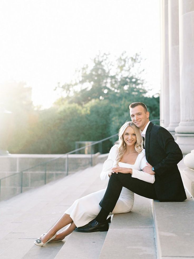 a man and woman sitting on the ground in front of a building, posing for a photo