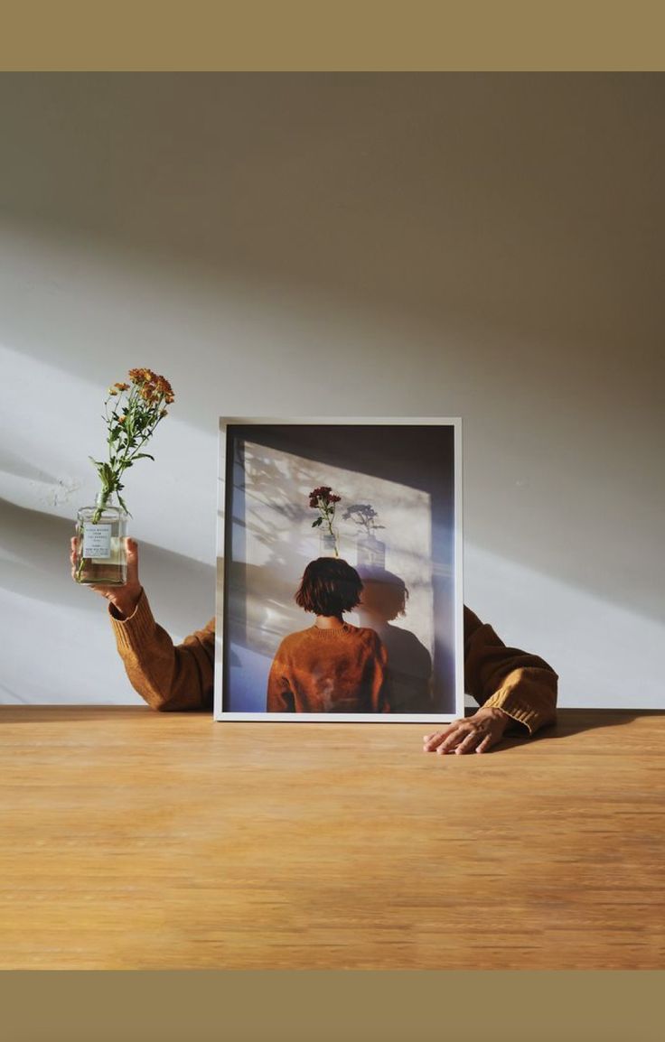 a person holding a vase with flowers in it on top of a wooden table next to a framed photograph