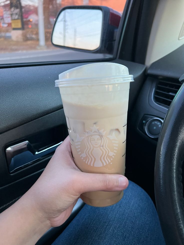 a hand holding a cup of coffee in front of a car steering wheel and dashboard