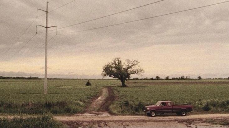 a red truck driving down a dirt road next to a tree and power lines on a cloudy day