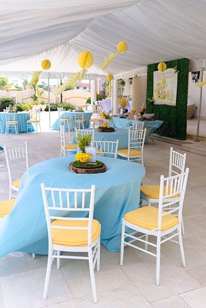 tables and chairs are set up in a tent for an outdoor event with blue tablecloths, yellow chair covers, and paper lanterns hanging from the ceiling