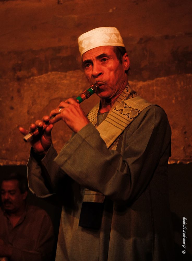 a man is playing the flute in front of other men and one has a white cap on his head