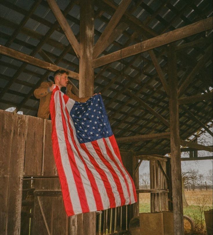 a man holding an american flag on top of a wooden structure in front of a barn