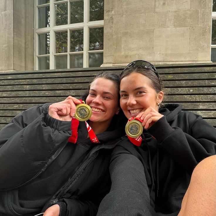 two women sitting on a bench with their medals in front of the building and stairs behind them